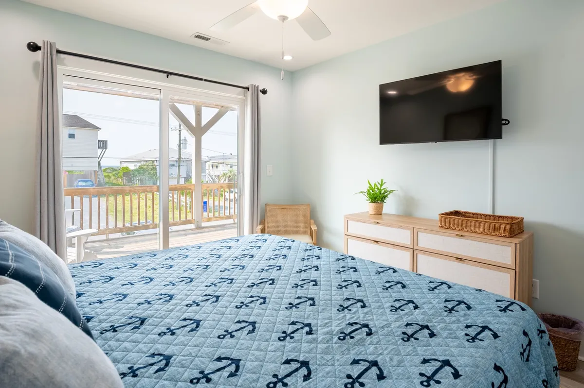 This image captures a serene bedroom with a bed dressed in an anchor-patterned quilt, a large window with sliding doors leading to a balcony, a wall-mounted smart TV, and a dresser with a small plant. The space feels bright, airy, and modern, with soft natural light and an ocean-inspired atmosphere.