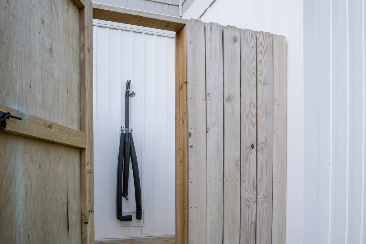 A simple but refreshing outdoor shower setup, with a shower hose attached to a wooden door and clean white walls, evokes the ease of post-beach clean-up and outdoor living.