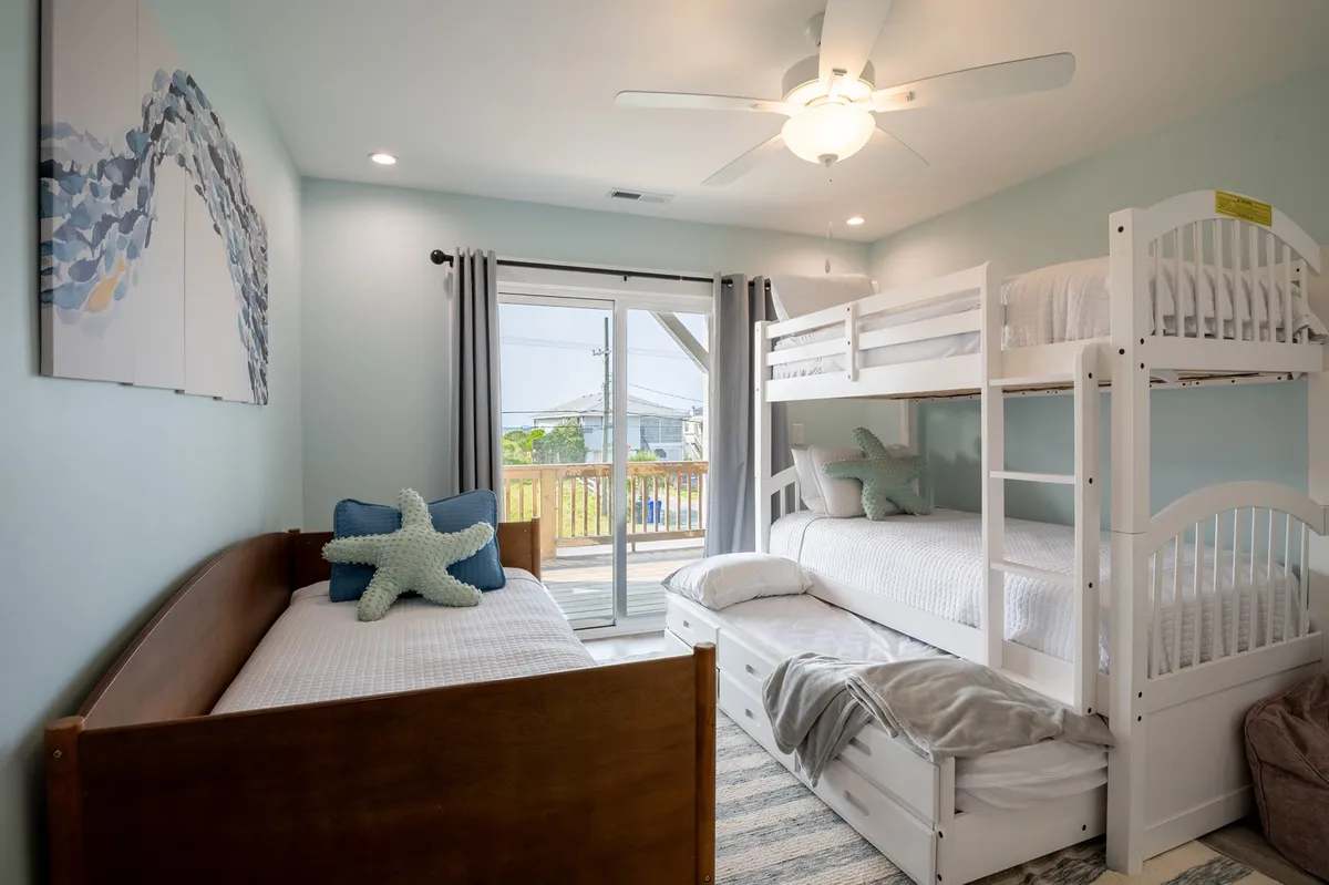 A playful, cozy bunk room featuring bunk beds, a day bed with a trundle, cheerful wall art, and a window letting in morning sunlight. The room is styled for children, with space for both play and rest.