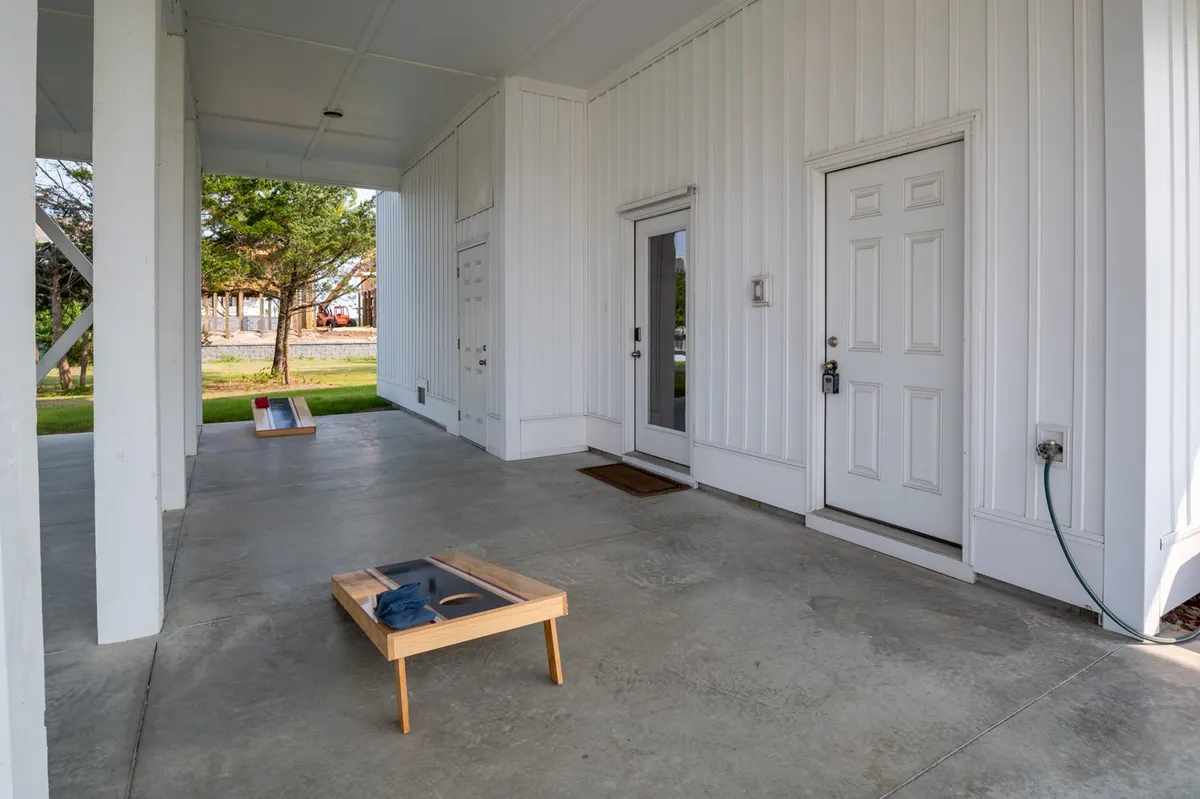 An inviting outdoor area featuring a cornhole game set up near the front doors, with a concrete floor and space for casual play and socializing. The scene suggests friendly family competitions and time spent outdoors.