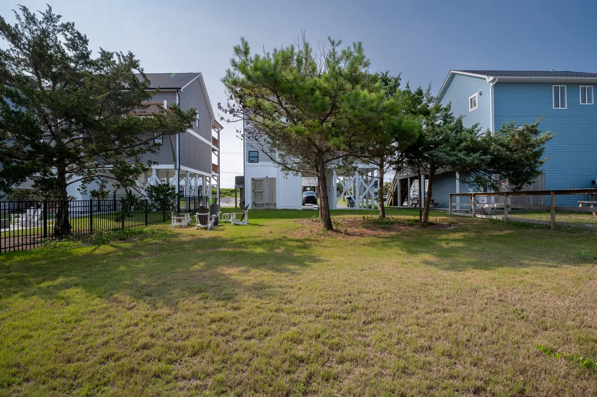A tranquil, sunlit open grassy yard surrounded by green trees and bordered by a fence, with nearby beach houses visible in the distance. The space offers a welcoming environment for outdoor activities, family picnics, and evening relaxation.