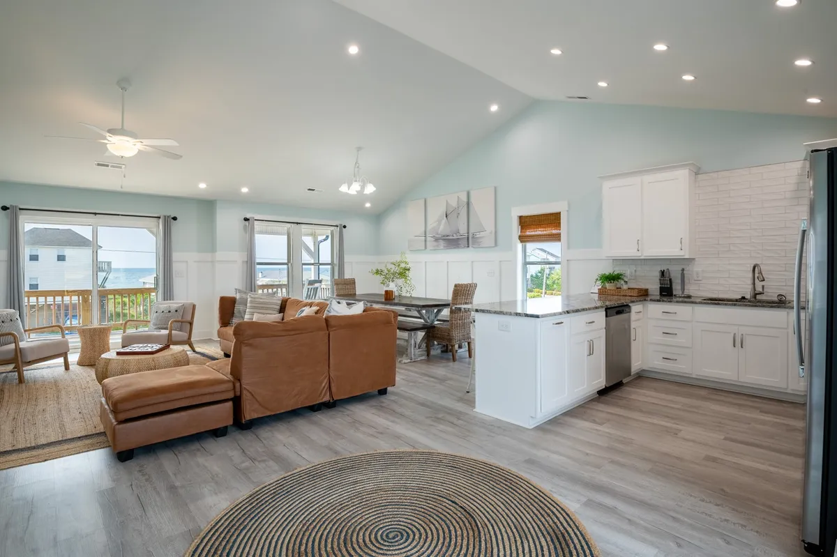 A spacious, light-filled living area featuring a large dining table, kitchen island, and expansive windows that frame beautiful views. The layout is designed for family gatherings, mealtime conversations, and shared moments.