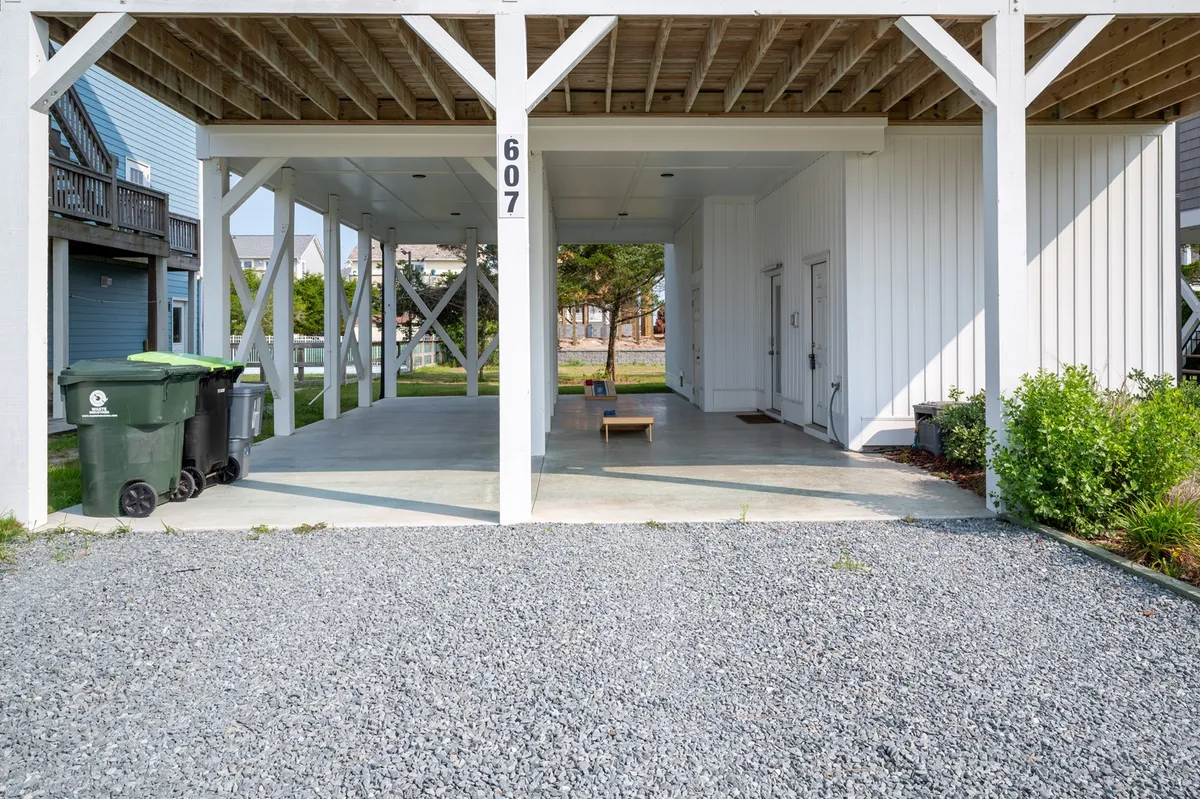 A ground-level view of the area beneath the house, featuring a bench for seating, convenient green trash bins for easy cleanup, and ample space for parking vehicles. The shaded, open area evokes a casual, welcoming atmosphere ideal for arrivals, unloading luggage, and informal gatherings.