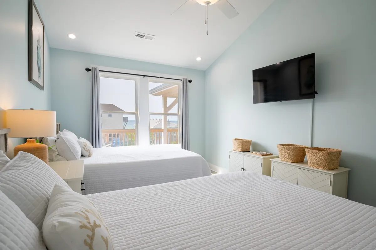 A spacious bedroom featuring two beds, large windows streaming in natural light, a wall-mounted TV, and basket storage. The decor is bright and comfortable, suitable for teens, couples, or extra guests.