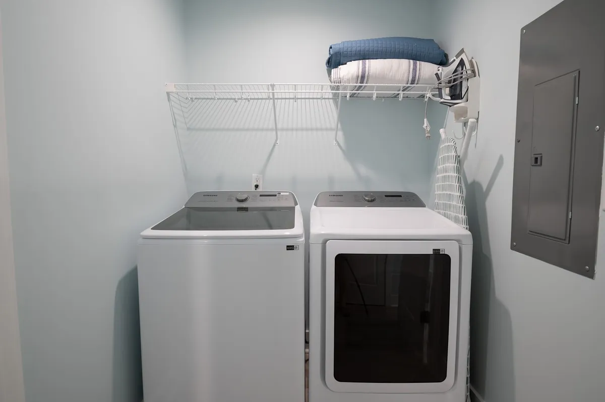 This image shows a well-organized laundry room with a washing machine, drying machine, an iron, an ironing board, and a shelf holding extra linens or a blanket. The setting is neat and practical, perfect for handling laundry after a day at the beach.