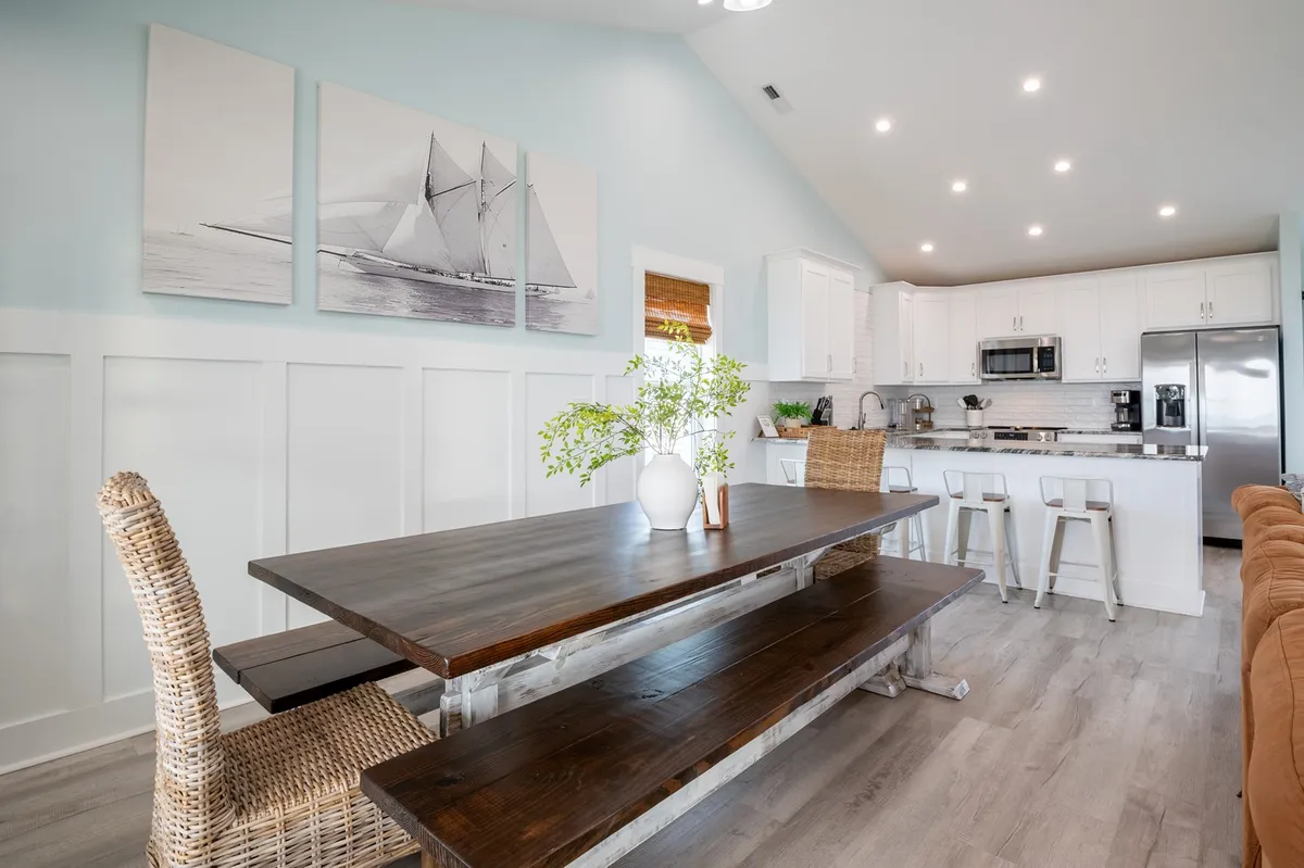 A spacious dining area features a large table, nautical-themed artwork, kitchen glimpses, and greenery in a vase. The setup is inviting for family meals and social gatherings, reflecting a relaxed coastal vibe.