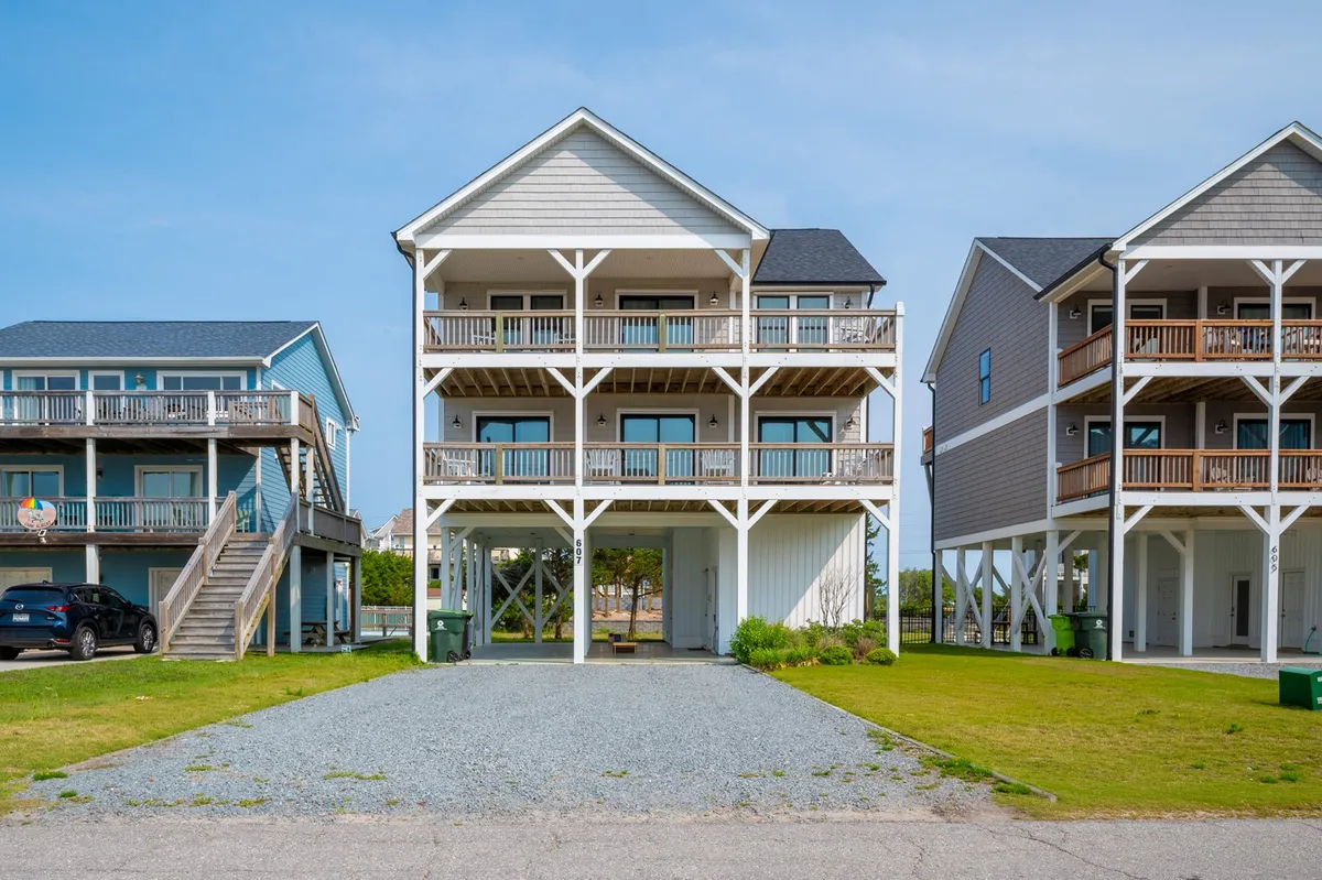 The hero image features a beautiful three-story beach house set against a backdrop of grassy dunes and neighboring homes. The home's spacious balconies offer sweeping ocean views, perfect for enjoying sunsets, morning coffee, and evenings under the stars. The inviting exterior hints at space for family gatherings and relaxed outdoor living.