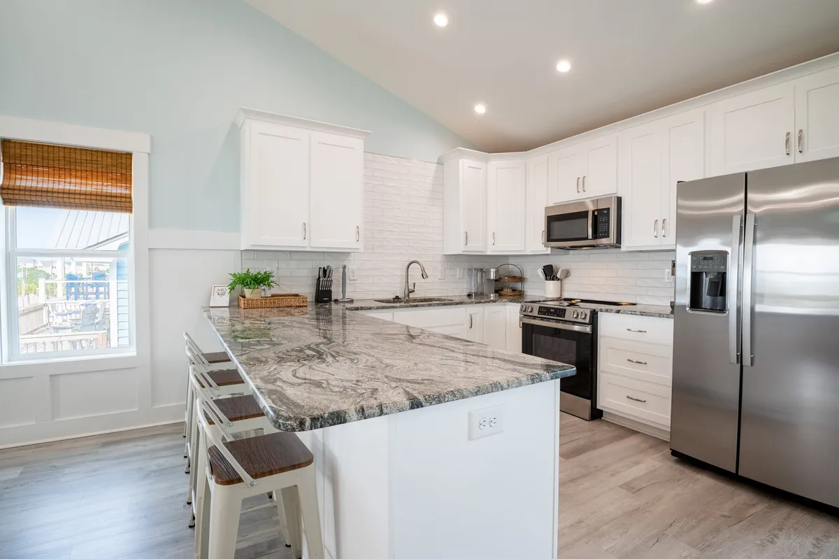 This interior shot showcases a modern, high-end kitchen with granite countertops, stainless steel appliances, and white cabinetry, bathed in natural light from a nearby window. The space is designed for both gourmet cooking and social gatherings.