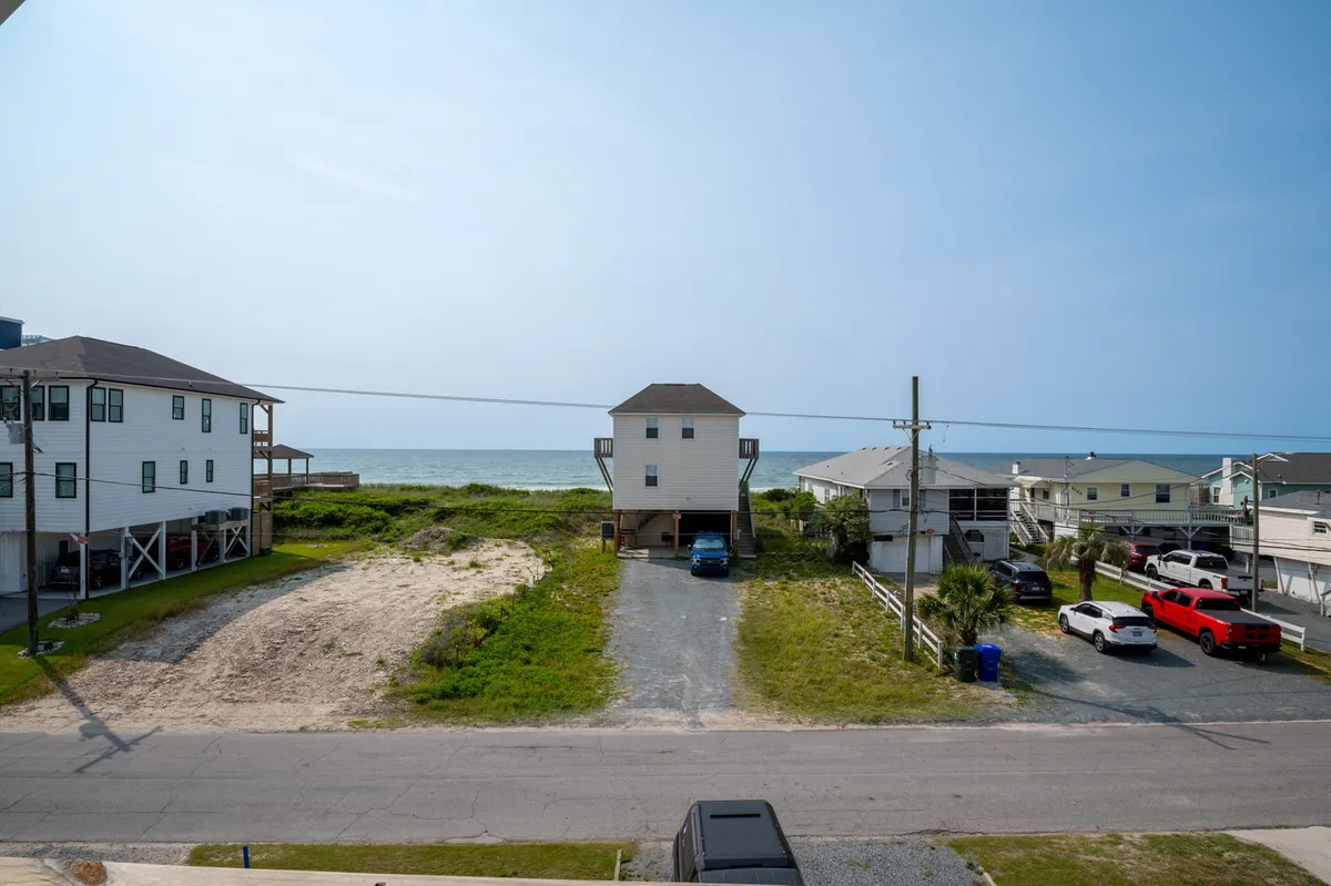 A wide view that encompasses the sandy beach, grassy areas, and surrounding beach houses. The image captures the easy beach access and the seamless transition from yard to shoreline. The mood is casual and welcoming, evoking the possibilities of beachcombing or evening walks.