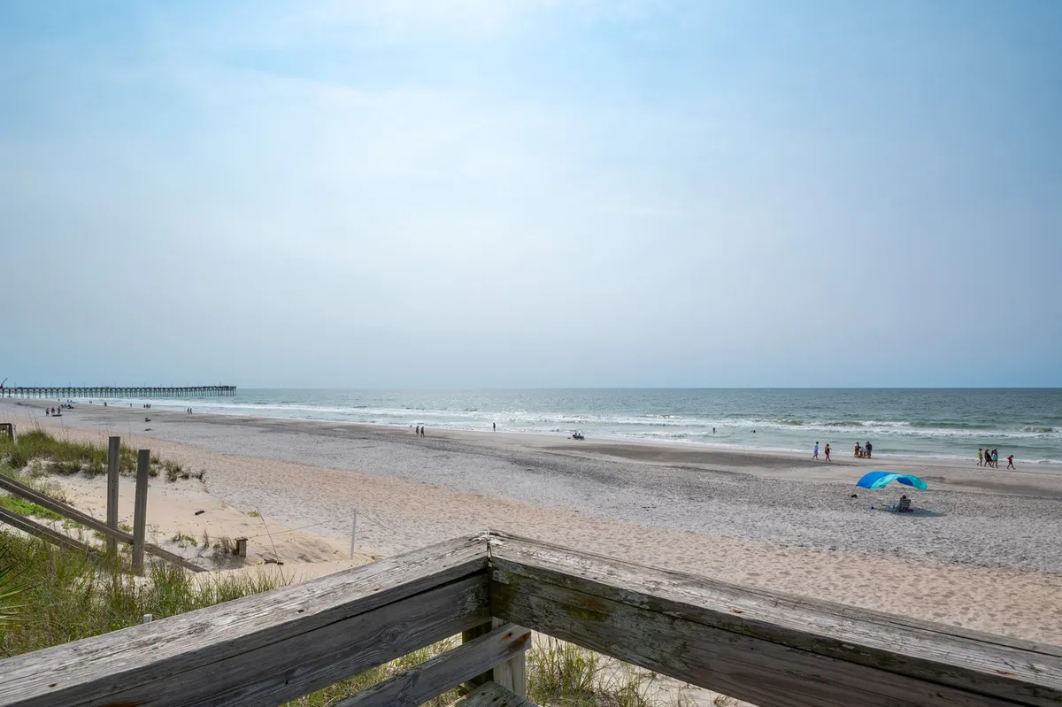 A wide, inviting view of the beach featuring the ocean, a pier, people, and a colorful umbrella. Families are seen swimming, strolling, sunbathing, and picnicking along the sandy shoreline. The scene is alive with activity yet retains a calming, tranquil vibe.