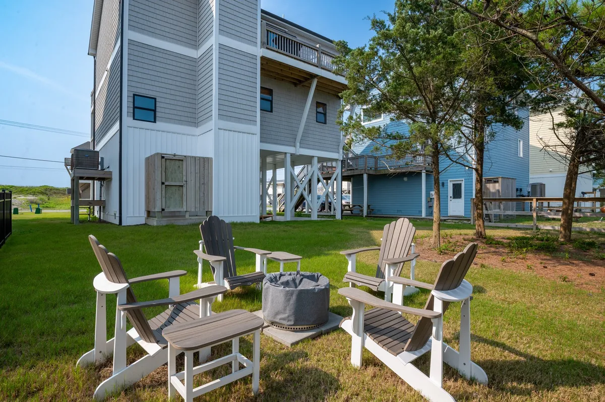 This image showcases a cozy fire pit area surrounded by Adirondack chairs, with a grassy yard and mature trees providing a lush, inviting backdrop. The exterior of the house is visible, reinforcing the connection between indoor and outdoor living. The setup is perfect for gatherings, storytelling, or roasting s'mores as described in the section.